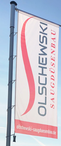 Biaa pionowa flaga na tle bkitnego nieba. Obrcone o 90 stopni logo firmy Olschewski Saugdsenbau e.K. jest na niej przedstawione.