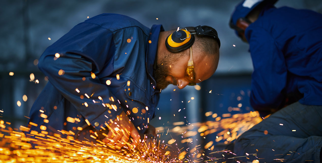 Zwei Männer in blauer Schutzkleidung mit Gehörschutz bearbeiten in einer Werkstatt Metall. Orange Funken sprühen.