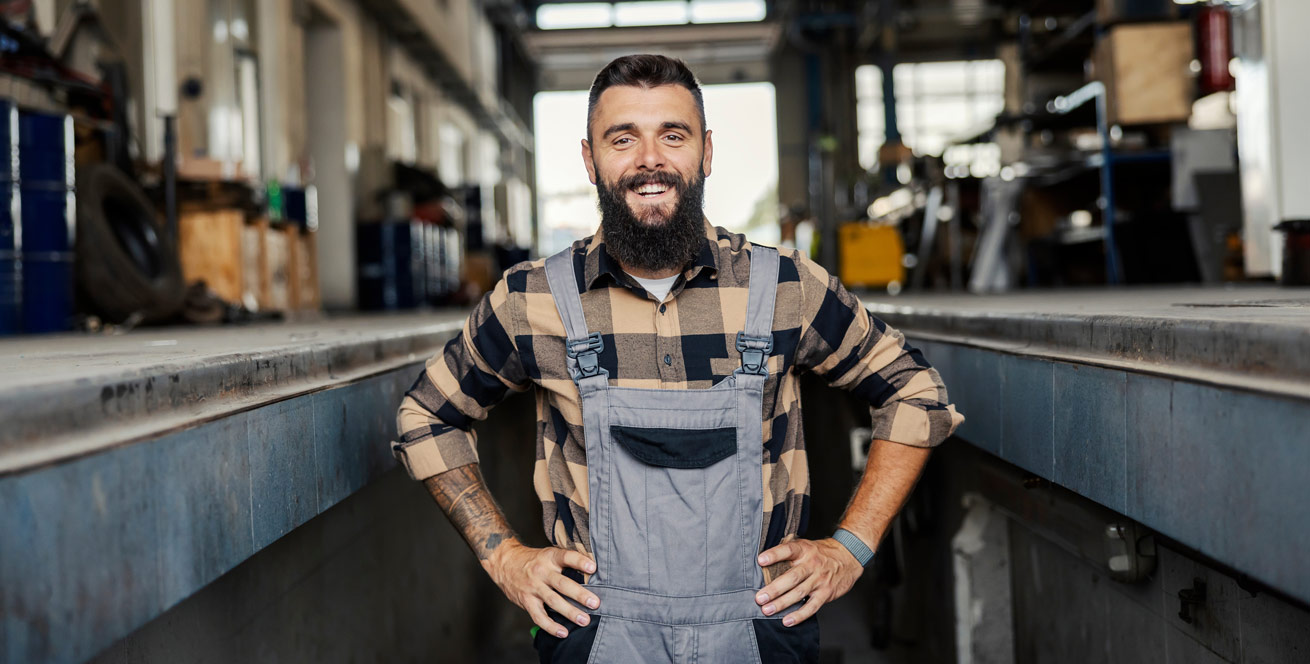 A man stands in work clothes smiling at the camera. He has his hands on his hips energetically. In the background you can see a metal construction workshop, tools and shelves.