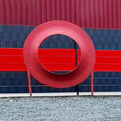 Frontaler Blick auf eine menschengroße rote Deckscheibe. Sie steht auf dunklem Schotter.Im Hintergrund ist ein grauer Zaun zu sehen über dessen gesamte Breite mittig ein roter handbreiter Streifen verläuft.